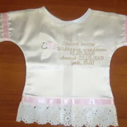 Biała, satynowa koszulka do chrztu z haftowanym napisem 'Chrzest Święty Wiktoria urodzona 13.02.2009 chrzest 23.05.2009 godz. 15.00', zdobiona koronką i różową wstążką, leżąca na drewnianym stole.