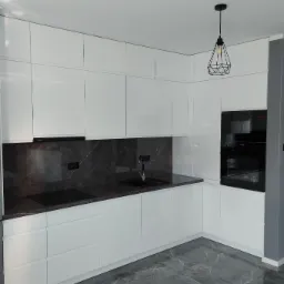 Biała, minimalistyczna kuchnia na wymiar z czarnym blatem imitującym marmur i geometryczną lampą wiszącą.