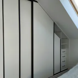 Biała szafa wnękowa z przesuwnymi drzwiami z czarnymi obramowaniami, dopasowana do skosu poddasza, z otwartą częścią z półkami i szufladami, częściowo zakryta folią ochronną.