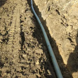 Długa, karbowana rura ułożona w wąskim wykopie w ziemi, widoczne ślady opon obok wykopu.