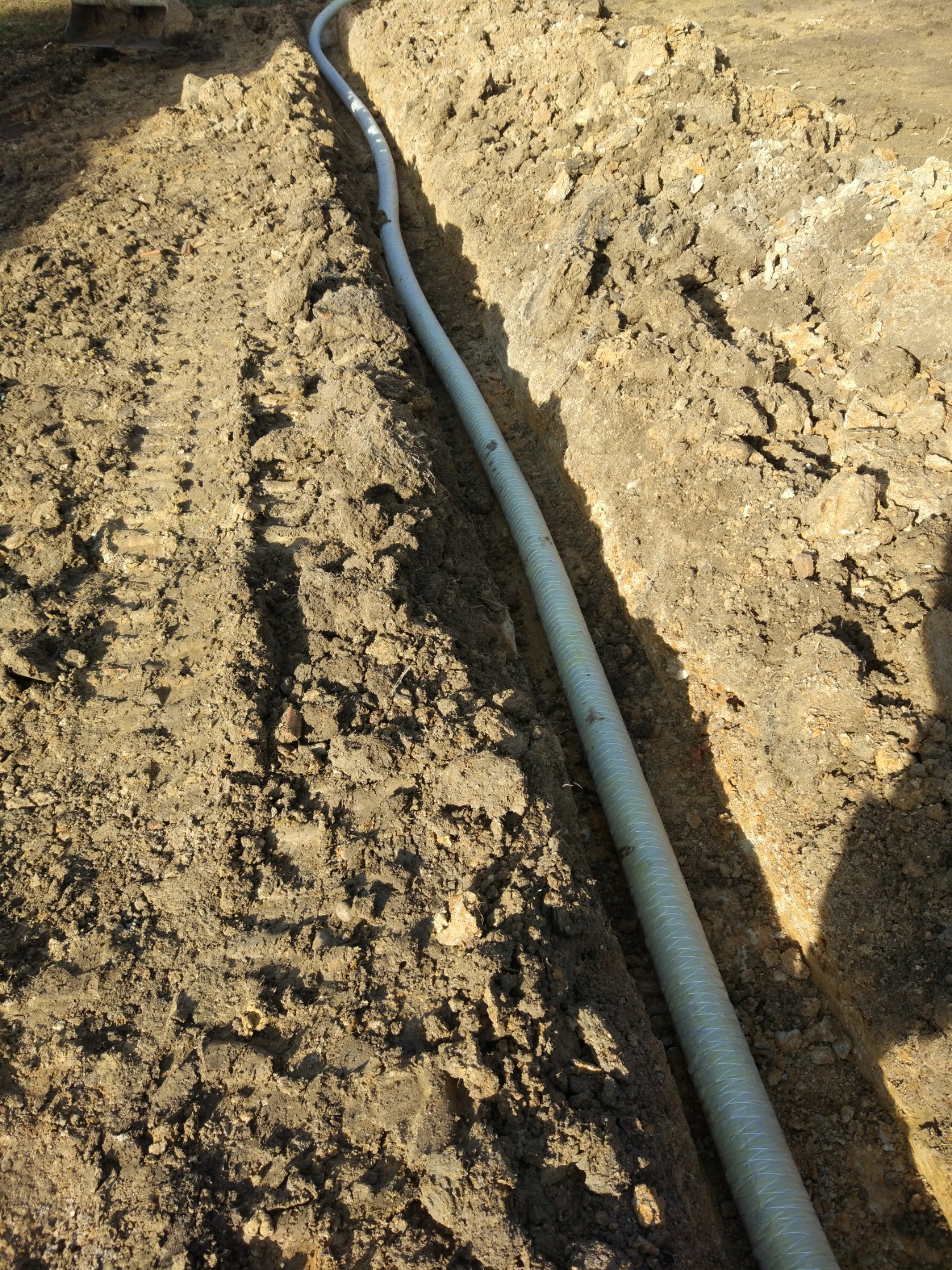 Długa, karbowana rura ułożona w wąskim wykopie w ziemi, widoczne ślady opon obok wykopu.