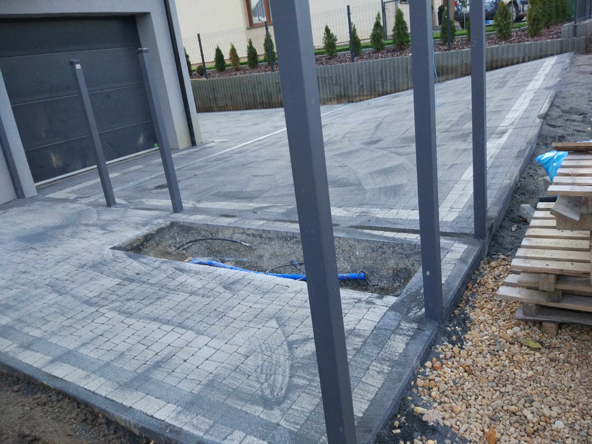 Fragment podjazdu z kostki brukowej w trakcie budowy pergoli, widoczne słupy konstrukcyjne, odsłonięte kable w wykopie, paleta drewniana obok.