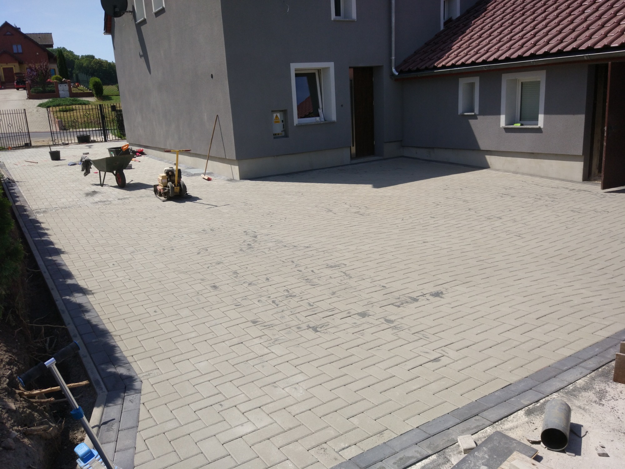 Nowa, jasnoszara kostka brukowa ułożona w jodełkę na podjeździe przed domem, widoczna taczka, ubijarka i narzędzia.