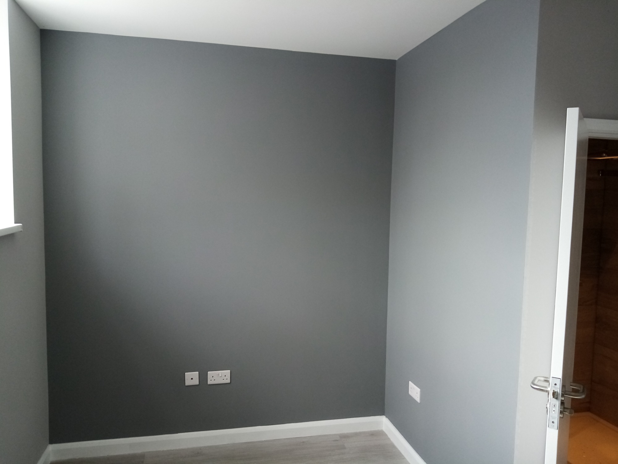 Pokój po malowaniu ścian na szaro, widoczne gniazdka elektryczne, listwy przypodłogowe i fragment otwartych drzwi do łazienki z drewnianym wykończeniem.