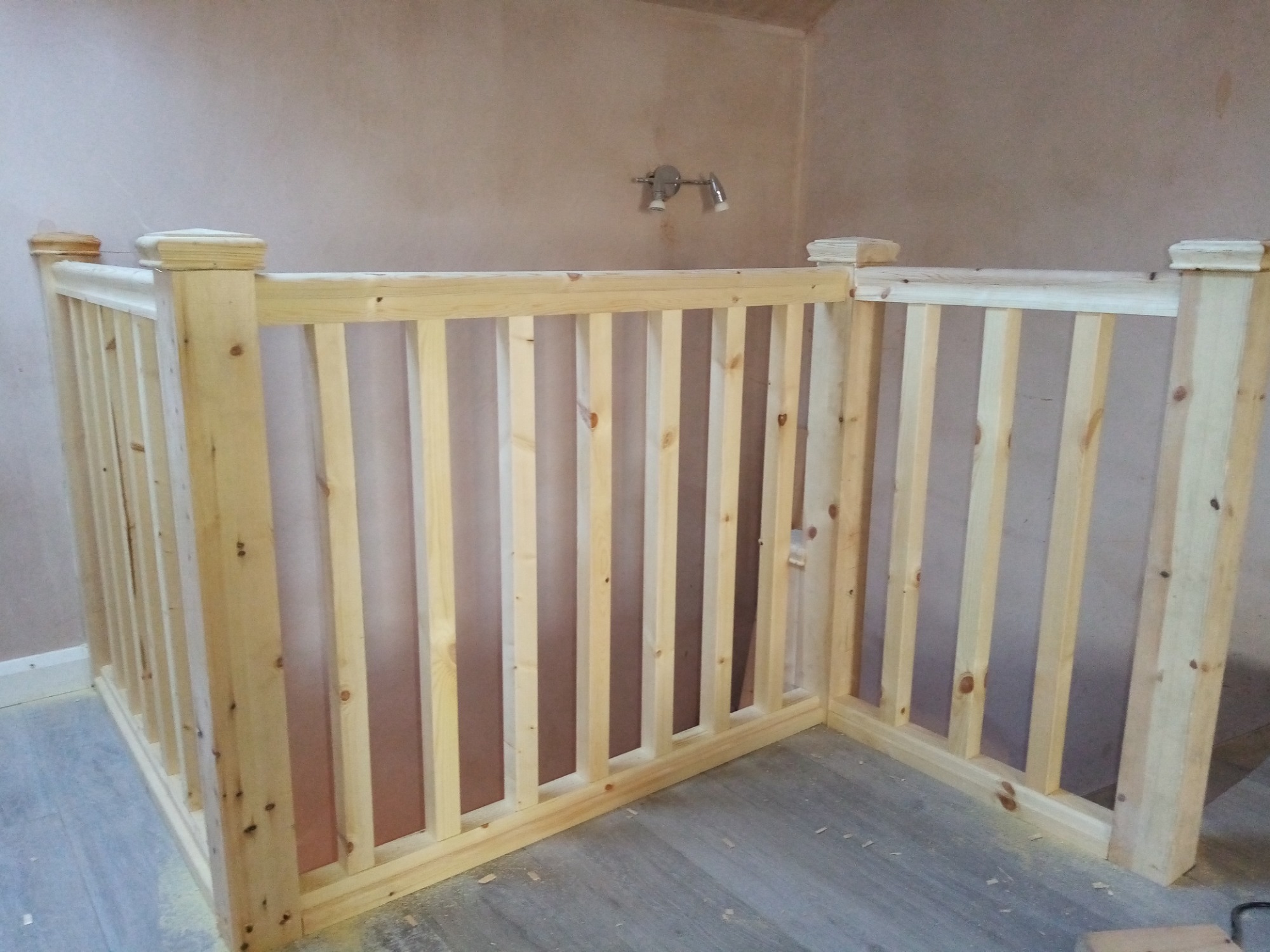 Surowa drewniana balustrada schodowa z prostymi szczeblami, zamontowana w rogu pokoju z surowymi ścianami i podwójnym reflektorem sufitowym.