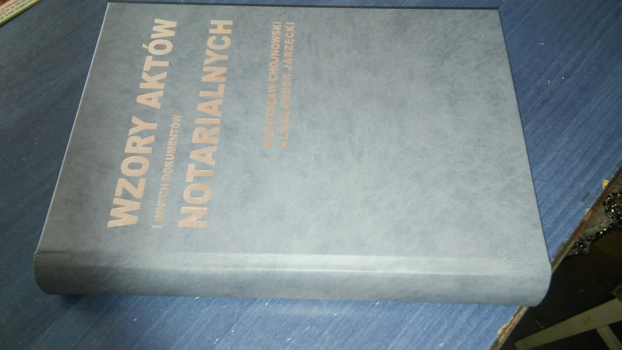 Książka w twardej oprawie z tytułem 'WZORY AKTÓW NOTARIALNYCH' w złotym kolorze, leżąca na niebieskim tle.
