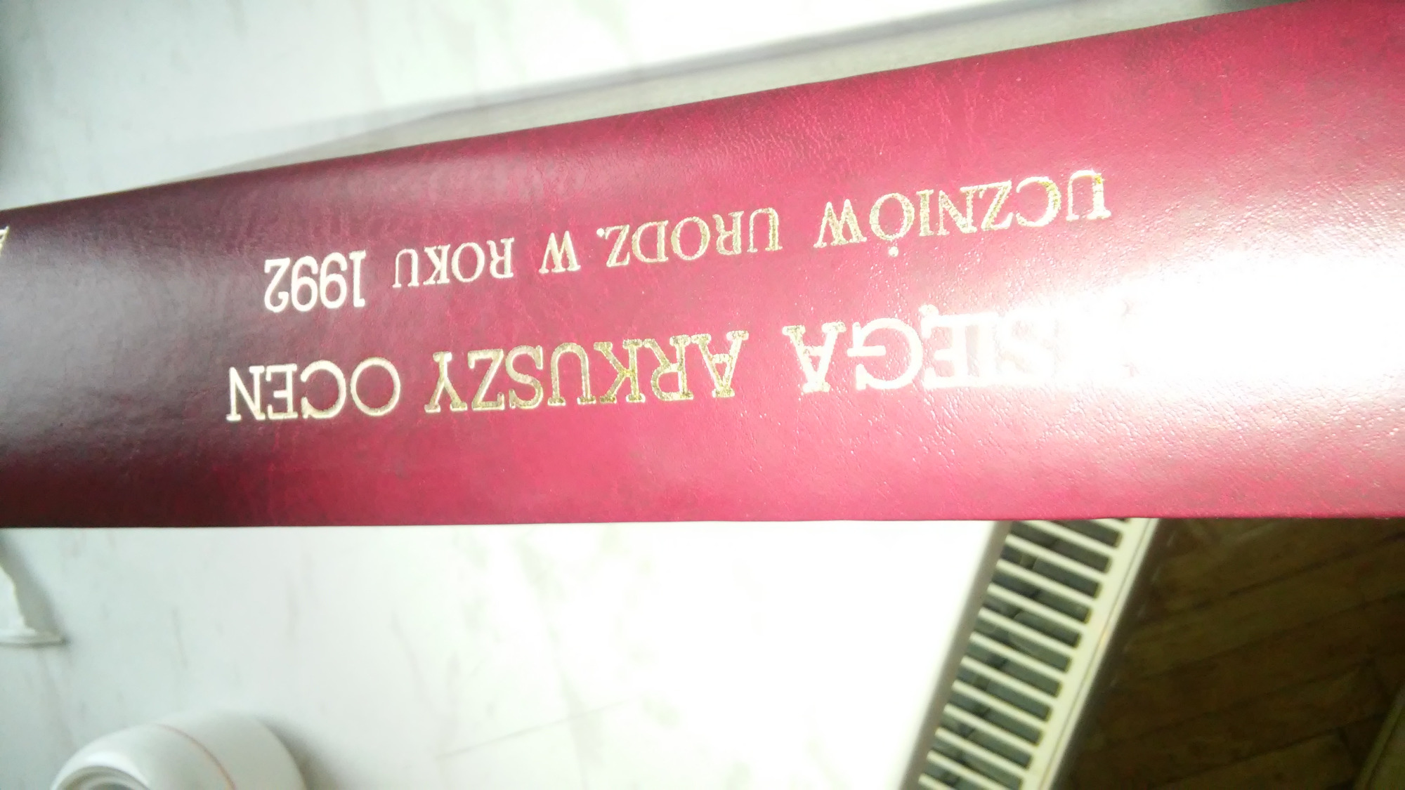 Czerwona oprawa książki z wytłoczonym złotym napisem 'KSIĘGA ARKUSZY OCEN UCZNIÓW URODZ. W ROKU 1992', widoczny fragment grzejnika.