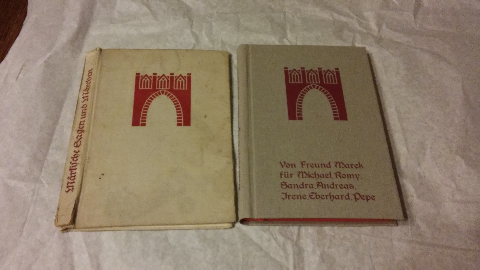 Dwie stare książki w twardej oprawie leżące obok siebie na pomarszczonym białym tle. Okładki zdobi czerwony rysunek bramy. Jedna książka ma tytuł na grzbiecie: Märkische Sagen und Märchen, a druga...