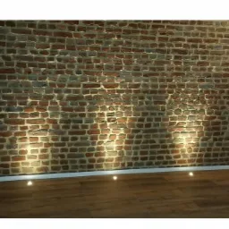 Ceglana ściana z oświetleniem podłogowym punktowym, widoczne gniazdko elektryczne i fragment podłogi z paneli.