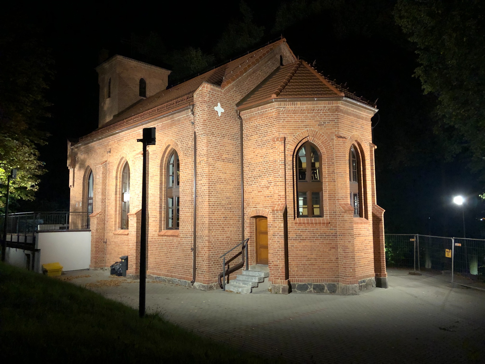 Oświetlony ceglany budynek z wieżyczką w nocy, widoczne okna z ciemnymi ramami i wejście ze schodami.