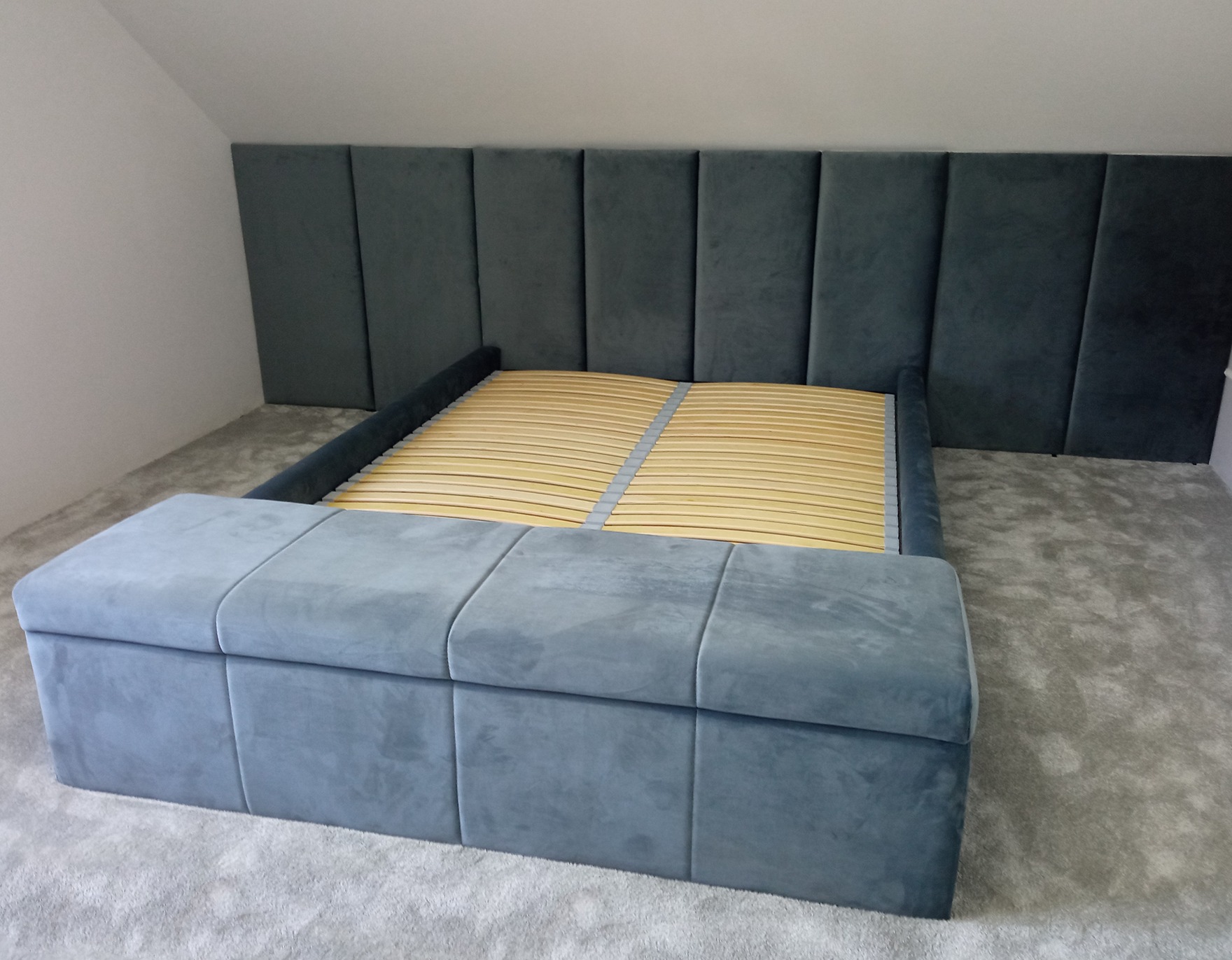 Tapicerowane łóżko z wysokim wezgłowiem w odcieniach szarości, z drewnianym stelażem i tapicerowanym pojemnikiem na pościel u podstawy, ustawione na szarym dywanie w pokoju na poddaszu.