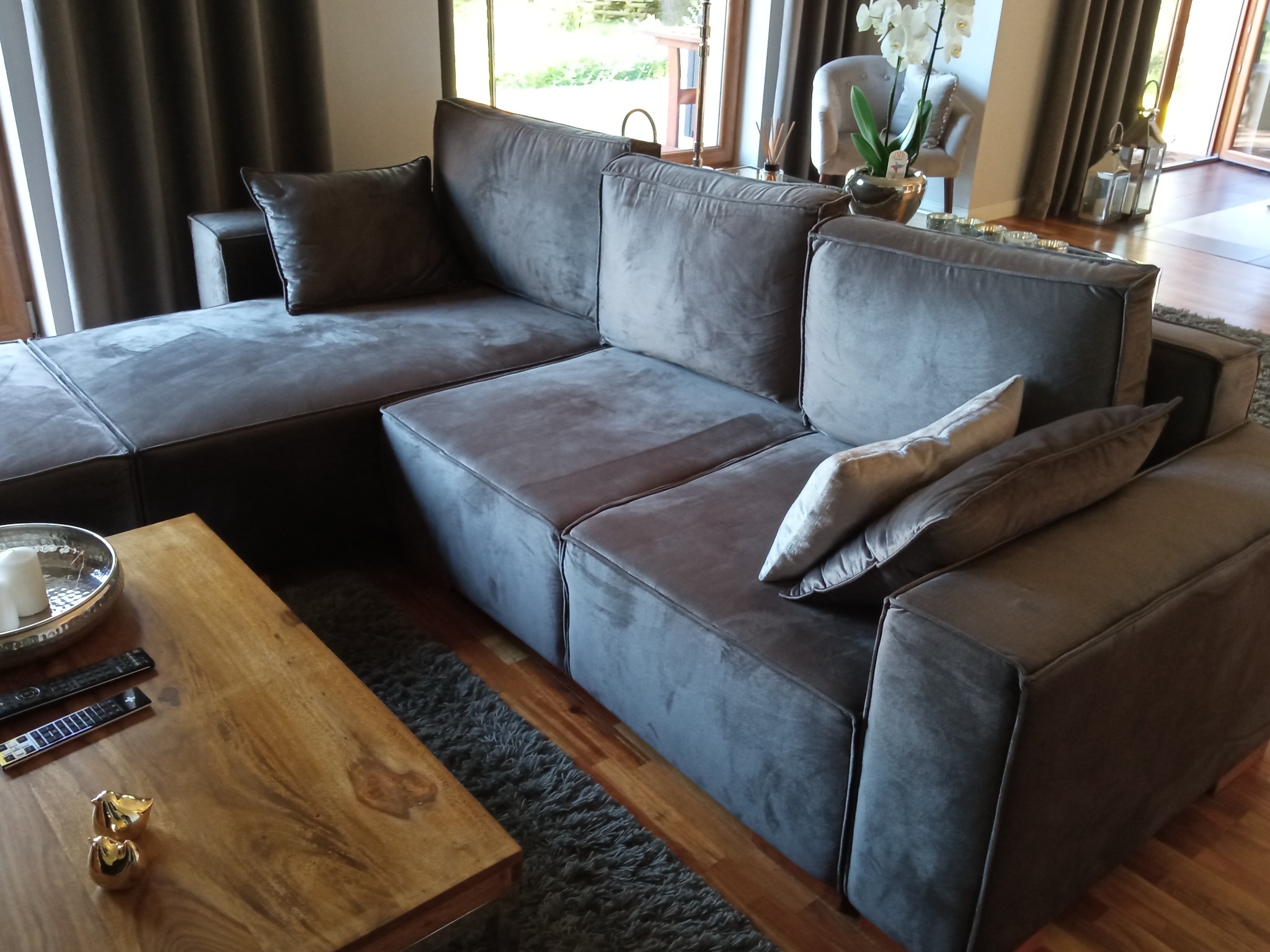 Szary, modułowy narożnik z weluru w salonie, widok z boku na drewniany stolik kawowy z pilotami i złotymi figurkami ptaków, w tle kwiaty i jasne krzesło.