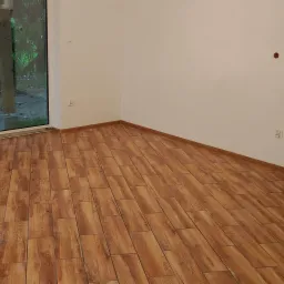 Wykończone wnętrze z podłogą wyłożoną panelami imitującymi drewno, widok na okno balkonowe z zielenią na zewnątrz, białe ściany z gniazdkami i włącznikami.