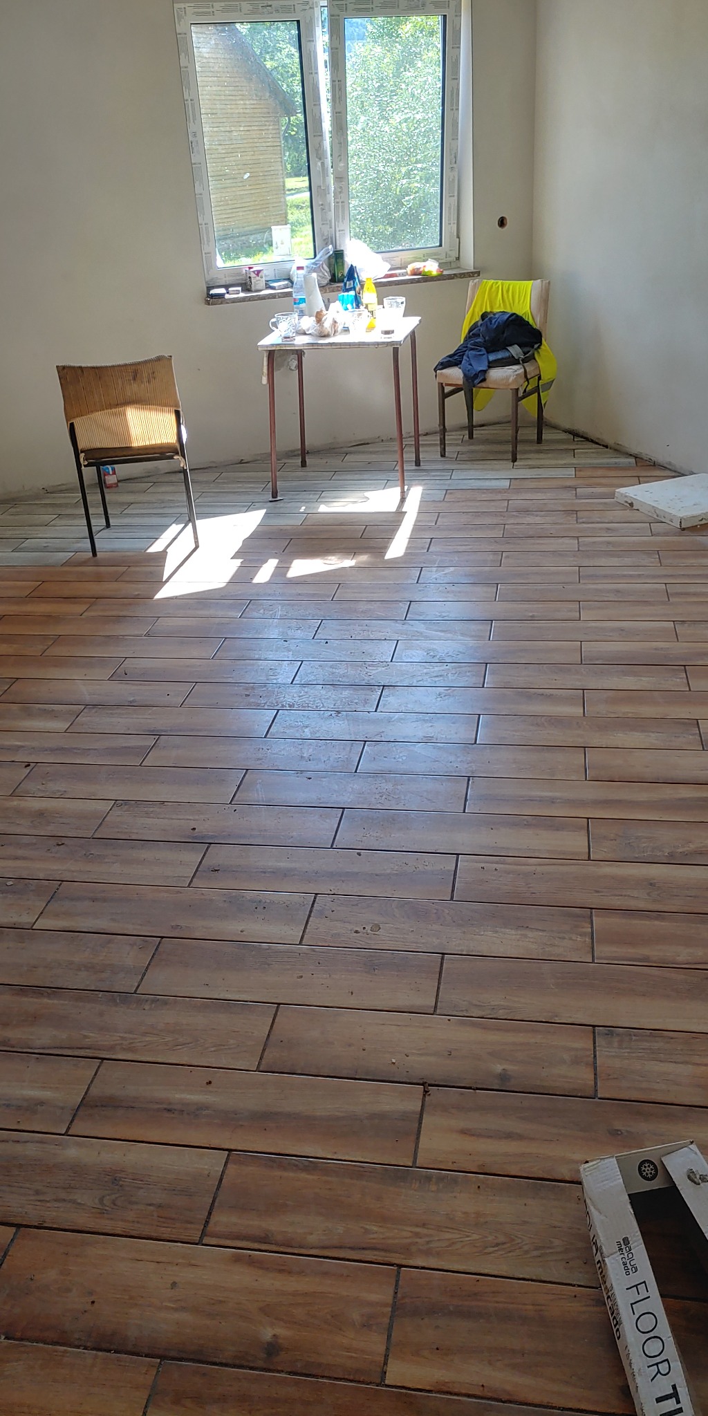 Ułożone brązowe płytki podłogowe imitujące drewno w słabo oświetlonym pokoju z oknem, stolikiem i krzesłami w tle. Częściowo widoczne kartony z płytkami w rogu.