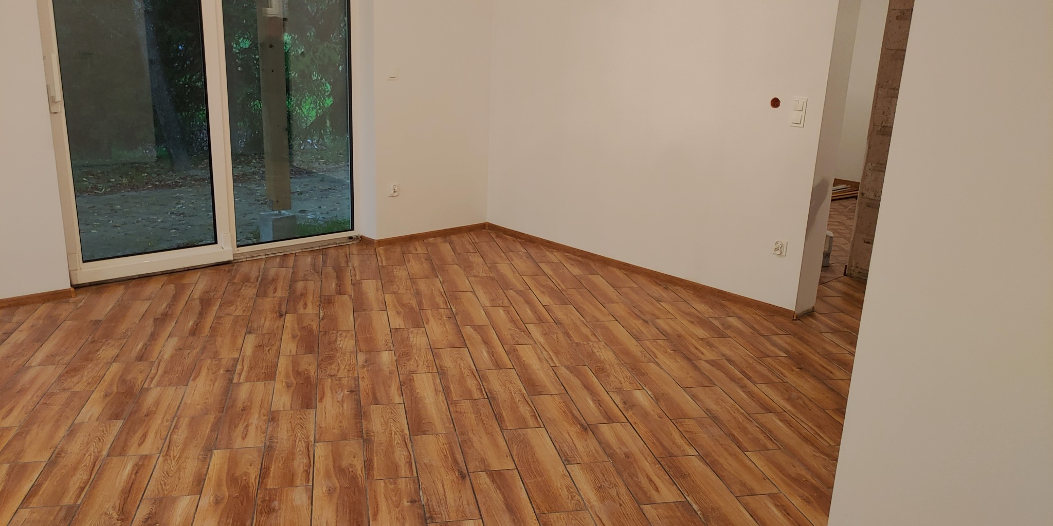 Wykończone wnętrze z podłogą wyłożoną panelami imitującymi drewno, widok na okno balkonowe z zielenią na zewnątrz, białe ściany z gniazdkami i włącznikami.