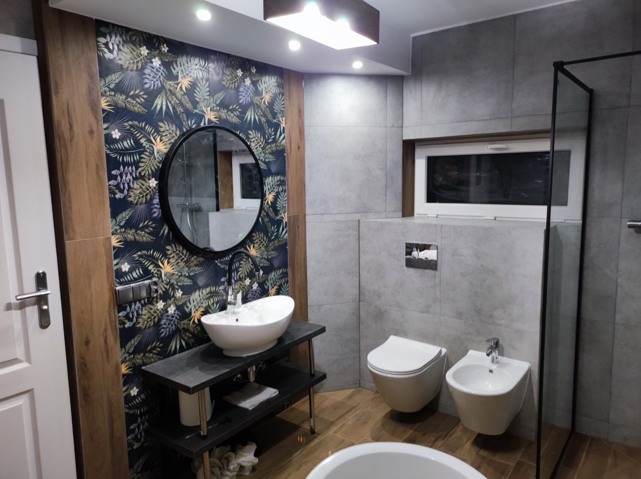 Nowoczesna łazienka z umywalką nablatową, okrągłym lustrem na ścianie z motywem roślinnym i szarą płytką imitującą beton, wisząca toaleta i bidet.