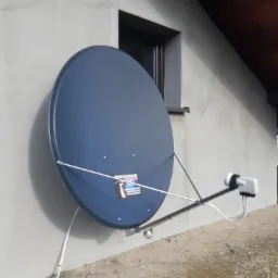 Niebieska antena satelitarna zamontowana na szarej ścianie budynku z widocznym oknem i fragmentem dachu, kable prowadzone wzdłuż ściany.