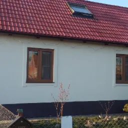 Dom jednorodzinny po tynkowaniu elewacji, z widocznymi brązowymi oknami, czerwoną dachówką i anteną satelitarną na ścianie bocznej.