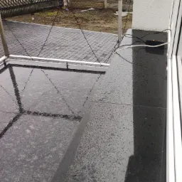 Granitowy parapet zewnętrzny po deszczu, widoczne krople wody, w tle taras wyłożony kostką i ogrodzenie.