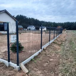 Nowo postawione, czarne ogrodzenie panelowe z betonową podmurówką otaczające posesję z domem jednorodzinnym, z małą choinką ozdobioną bombkami przy jednym ze słupków.