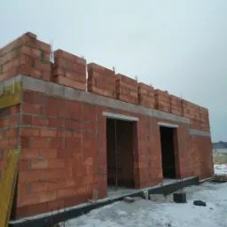 Budowa domu jednorodzinnego w stanie surowym otwartym, widoczne ściany z czerwonej cegły i betonowe fundamenty w zimowej scenerii.