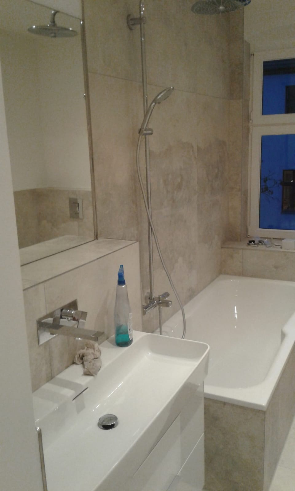 Nowoczesna łazienka z umywalką nablatową, chromowaną armaturą i wanną, wykończona płytkami w odcieniach beżu, z widocznym oknem i butelką płynu do czyszczenia.