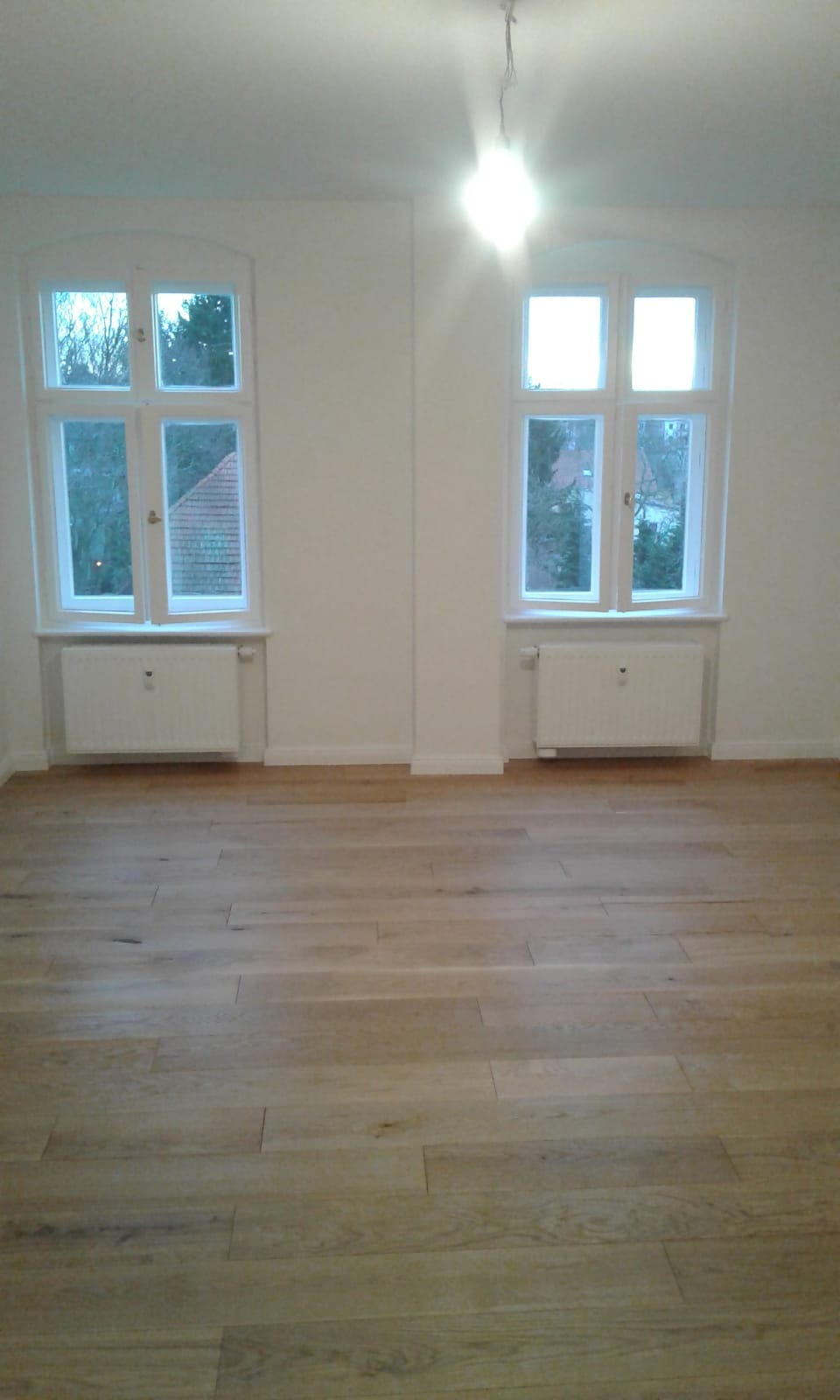 Jasne, dębowe deski podłogowe w pustym pokoju z dwoma oknami z białymi ramami i grzejnikami pod parapetami, widoczne okablowanie od sufitowej lampy.