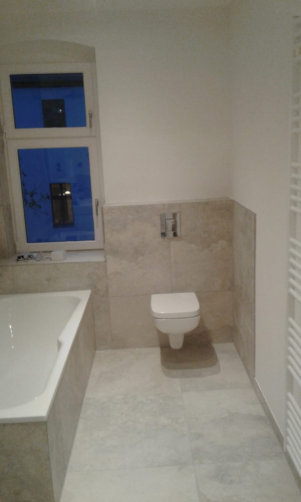 Nowoczesna łazienka z beżowymi płytkami, wisząca toaleta, biała wanna i grzejnik drabinkowy, widok na nocne niebo przez okno.