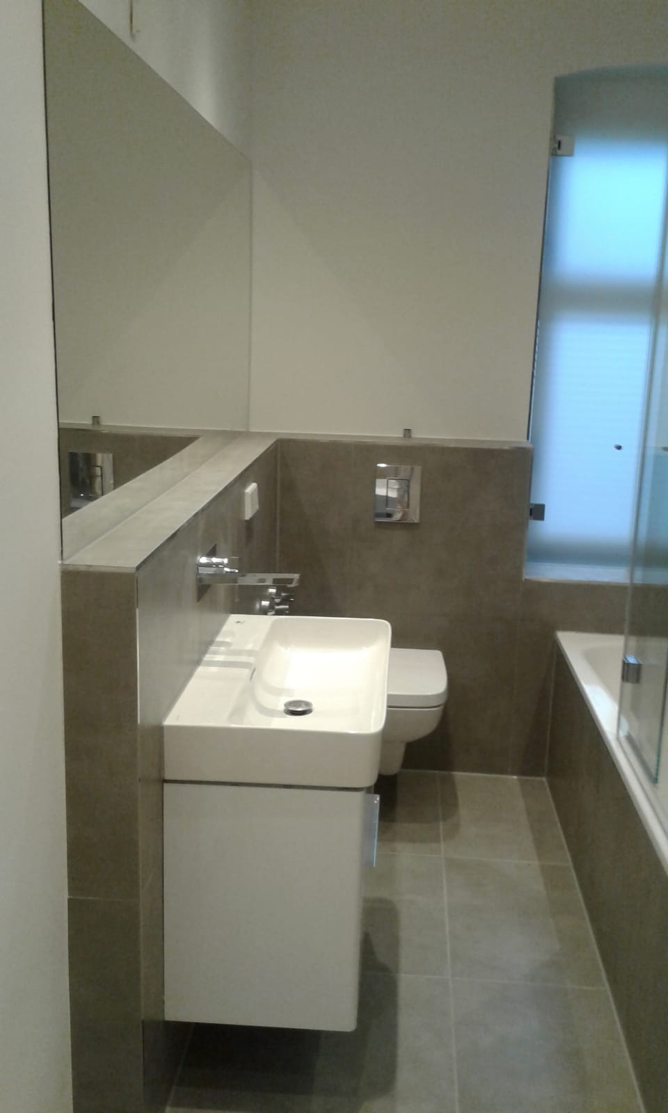 Nowoczesna łazienka z umywalką nablatową na szafce, dużym lustrem i szarą glazurą, widoczny fragment wanny z parawanem.