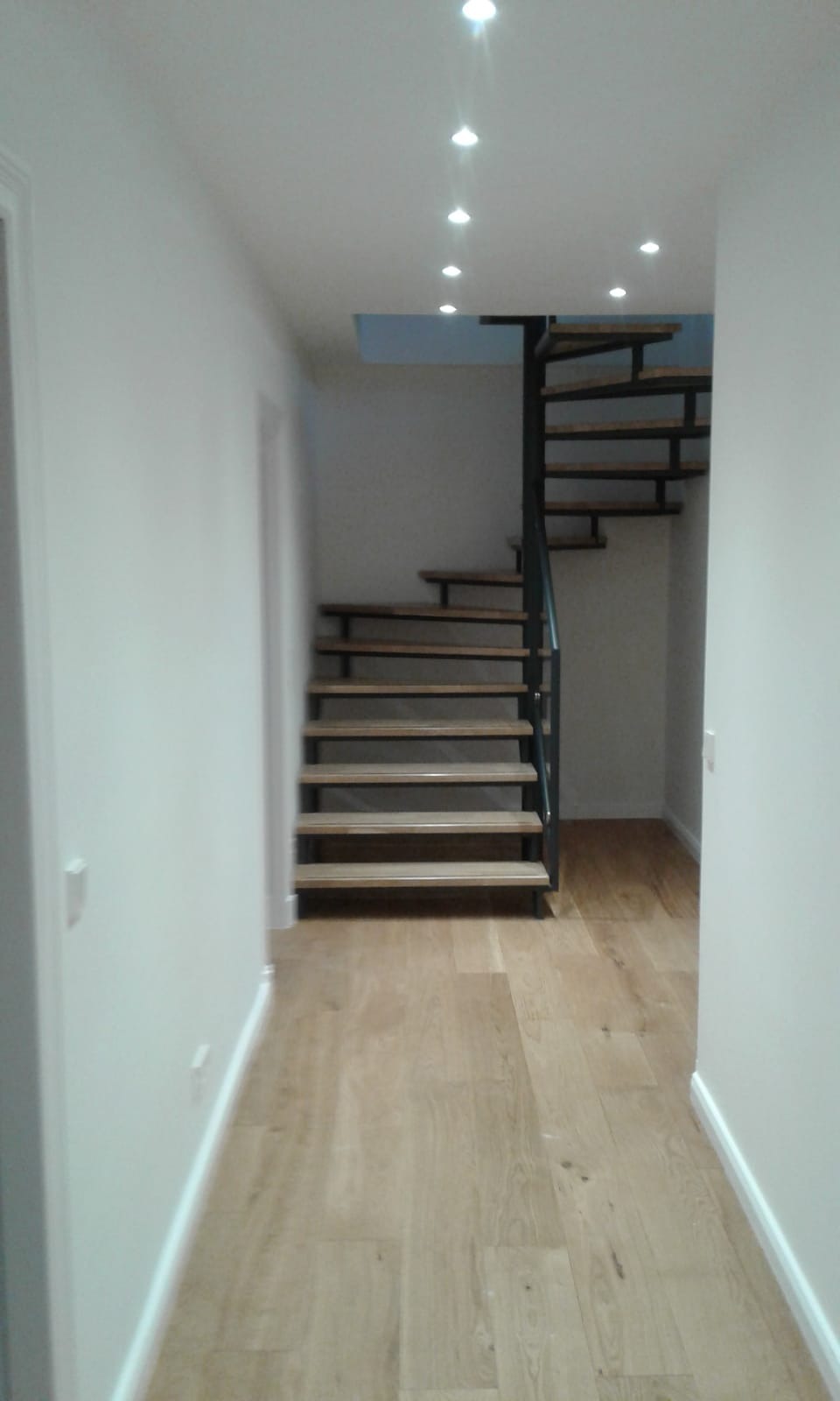 Wnętrze domu z drewnianą podłogą i spiralnymi schodami z drewnianymi stopniami i czarną metalową konstrukcją, oświetlone punktowymi lampami sufitowymi.