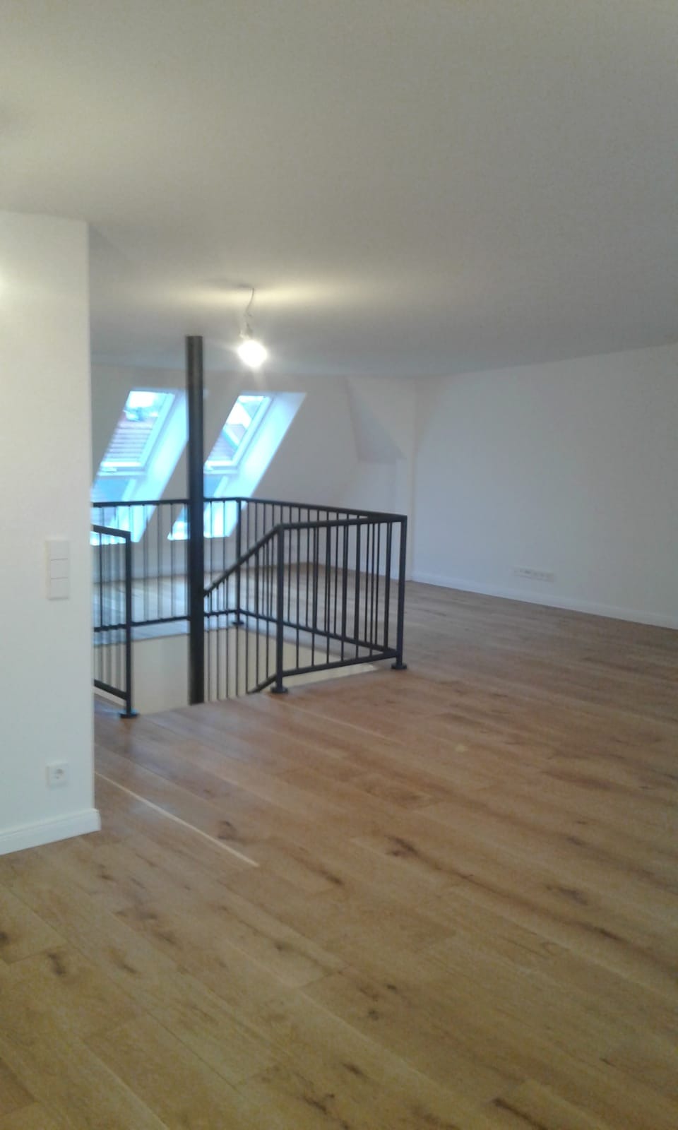 Jasne wnętrze poddasza z drewnianą podłogą, czarną balustradą schodów i oknami dachowymi w skosach dachu.