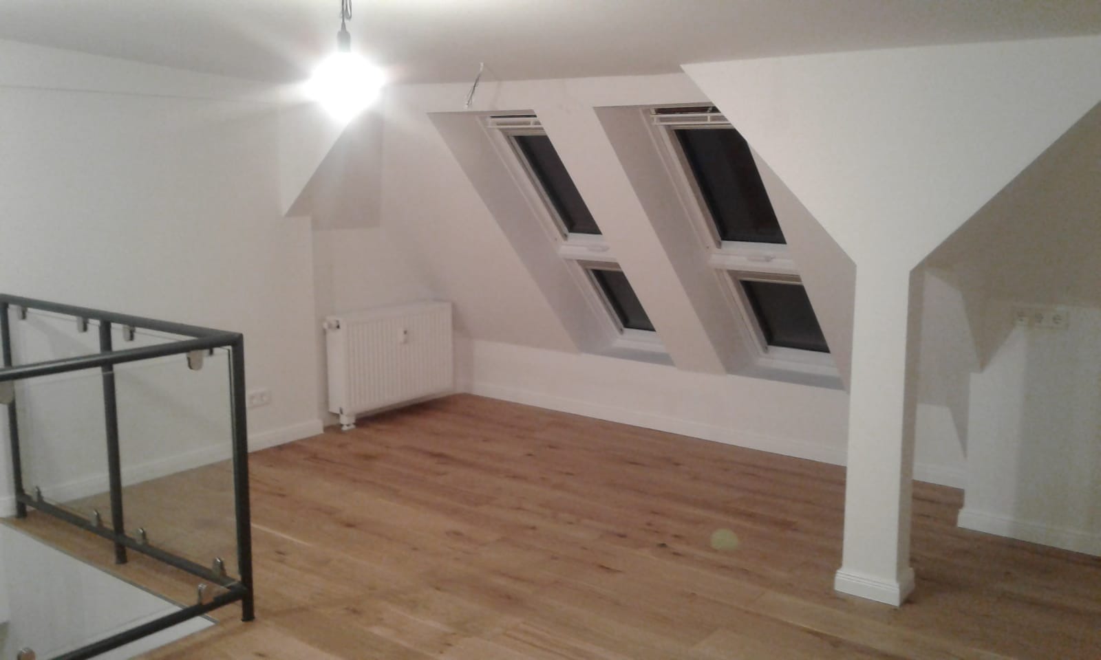 Jasne, przestronne poddasze po remoncie z drewnianą podłogą, dwoma oknami dachowymi w skosie, białymi ścianami i grzejnikiem. Widoczna balustrada ze szkła i metalu.