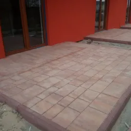 Ułożona kostka brukowa w kolorze ceglastym tworząca taras przy czerwonym budynku z drewnianymi oknami, widoczne obrzeża i piasek.