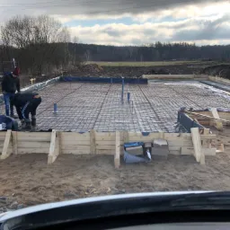 Przygotowanie zbrojenia i szalunku na płycie fundamentowej pod budowę domu, widoczni dwaj pracownicy w trakcie pracy, teren budowy z widokiem na las w tle.