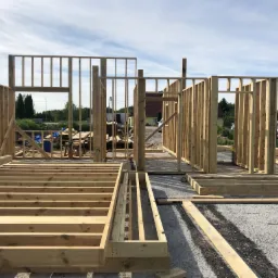 Wznoszenie konstrukcji szkieletowej domu jednorodzinnego z widocznymi elementami drewnianymi podłogi i ścian w słoneczny dzień.