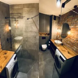 Industrialna łazienka z kabiną prysznicową typu walk-in, umywalką nablatową na drewnianym blacie, pralką i ścianą z cegły, oświetlona wiszącymi żarówkami.