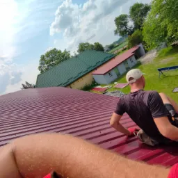 Montaż bordowej blachy trapezowej na dachu budynku gospodarczego, widok z perspektywy pracownika w czerwonych rękawicach, w tle budynki i zielone drzewa pod błękitnym, pochmurnym niebem.