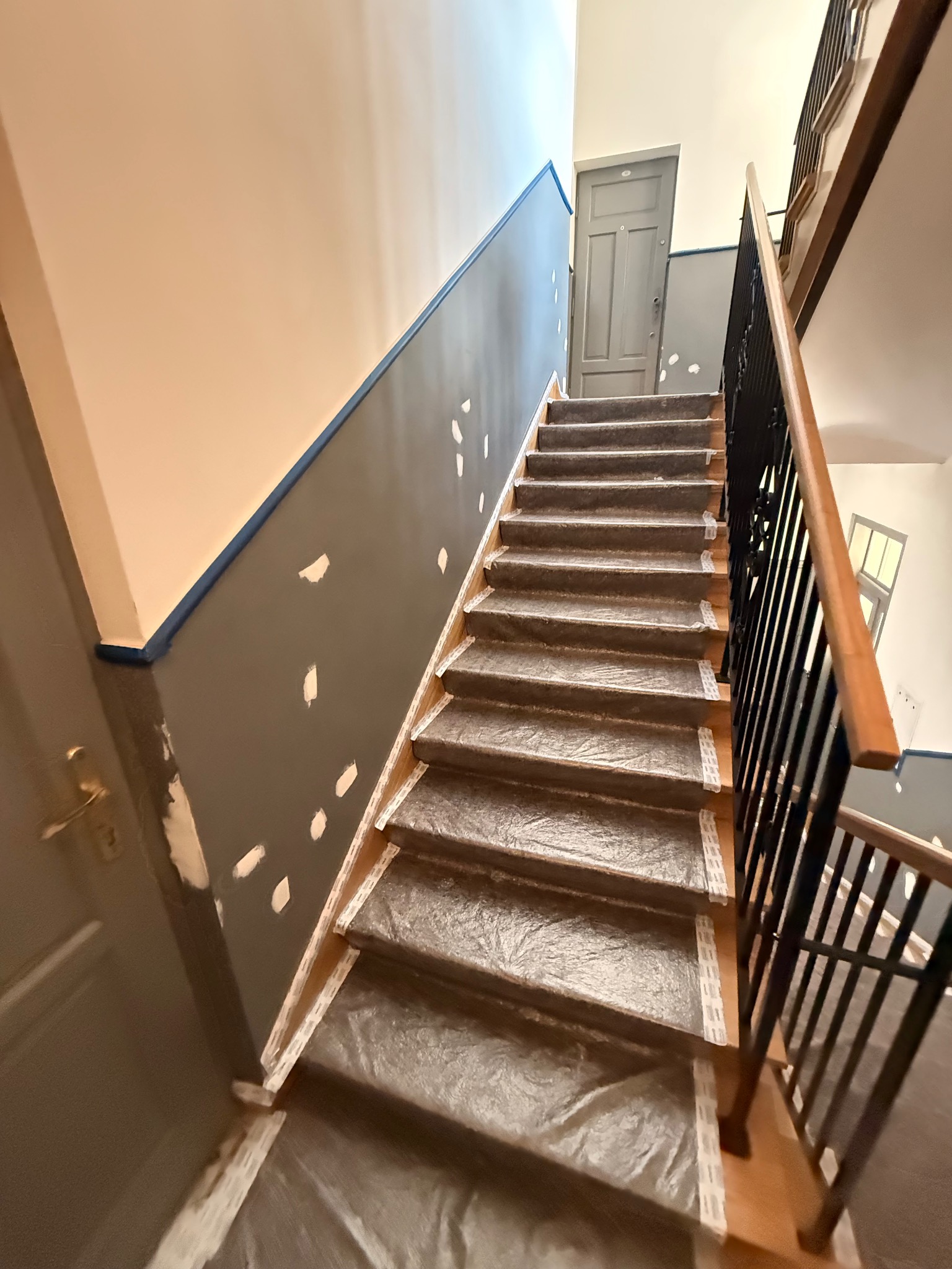 Klatka schodowa w trakcie malowania: ściany z szarą farbą, zabezpieczone taśmą i folią schody. Widoczne białe plamy po szpachlowaniu. Górne piętro z drzwiami.