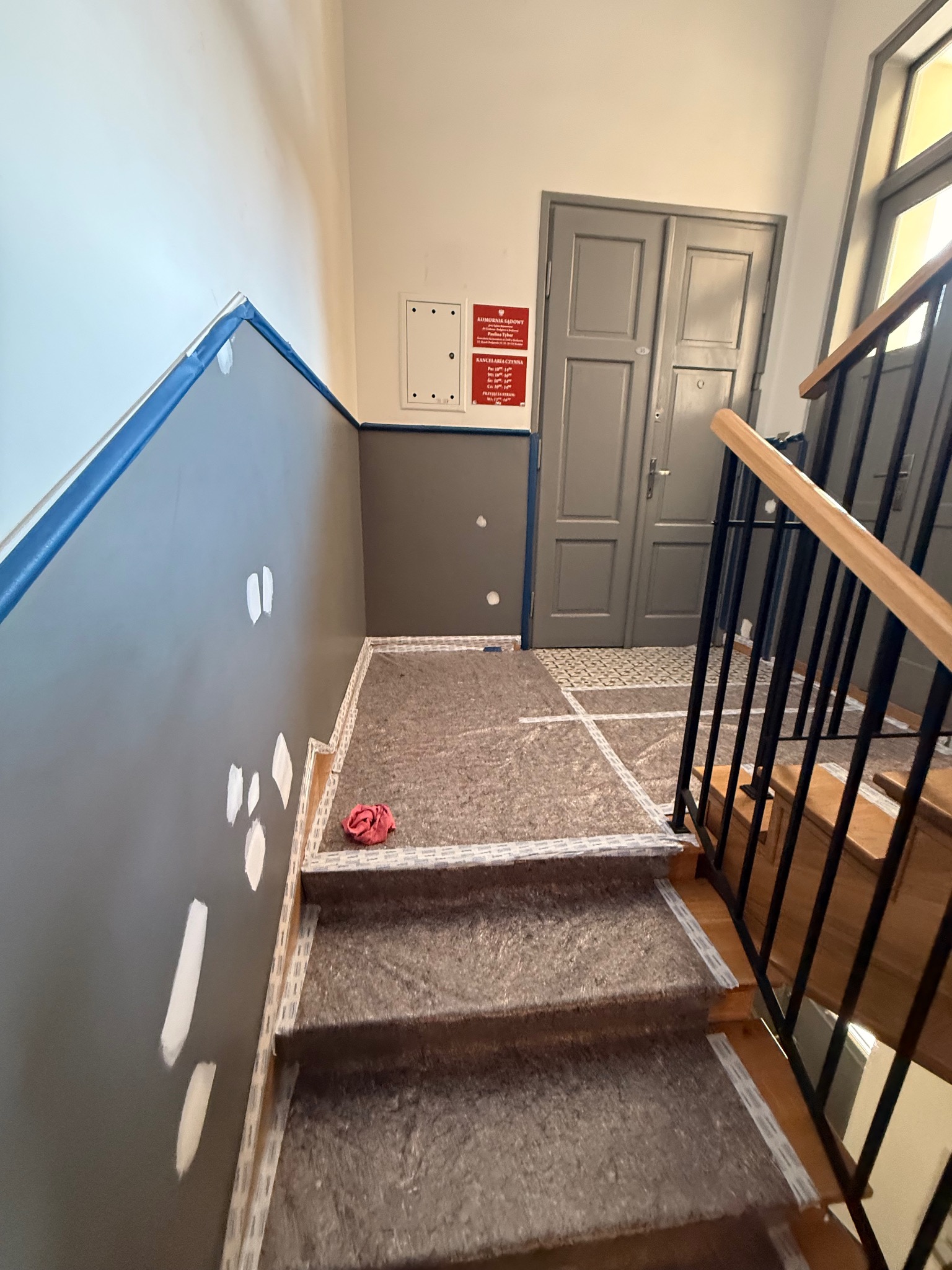 Klatka schodowa w trakcie malowania: szare ściany z białymi łatami, zabezpieczone schody i podłoga. Widoczne drzwi i fragment balustrady.