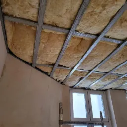 Montaż izolacji z wełny mineralnej w metalowym stelażu sufitu podwieszanego, widok na okno i fragment ściany w surowym stanie.