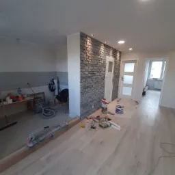 Wnętrze w trakcie prac wykończeniowych: położona podłoga z paneli, ściana z dekoracyjnej cegły z oświetleniem punktowym, widoczne narzędzia i materiały budowlane, w tle pomieszczenie...