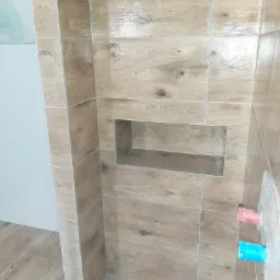 Ułożone płytki imitujące drewno w kabinie prysznicowej z wnęką, widoczne fragmenty instalacji wodnej i odpływ liniowy.