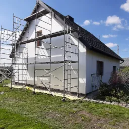 Dom jednorodzinny w trakcie ocieplania, z widocznymi rusztowaniami, styropianem na ścianach i pojazdem dostawczym w tle.