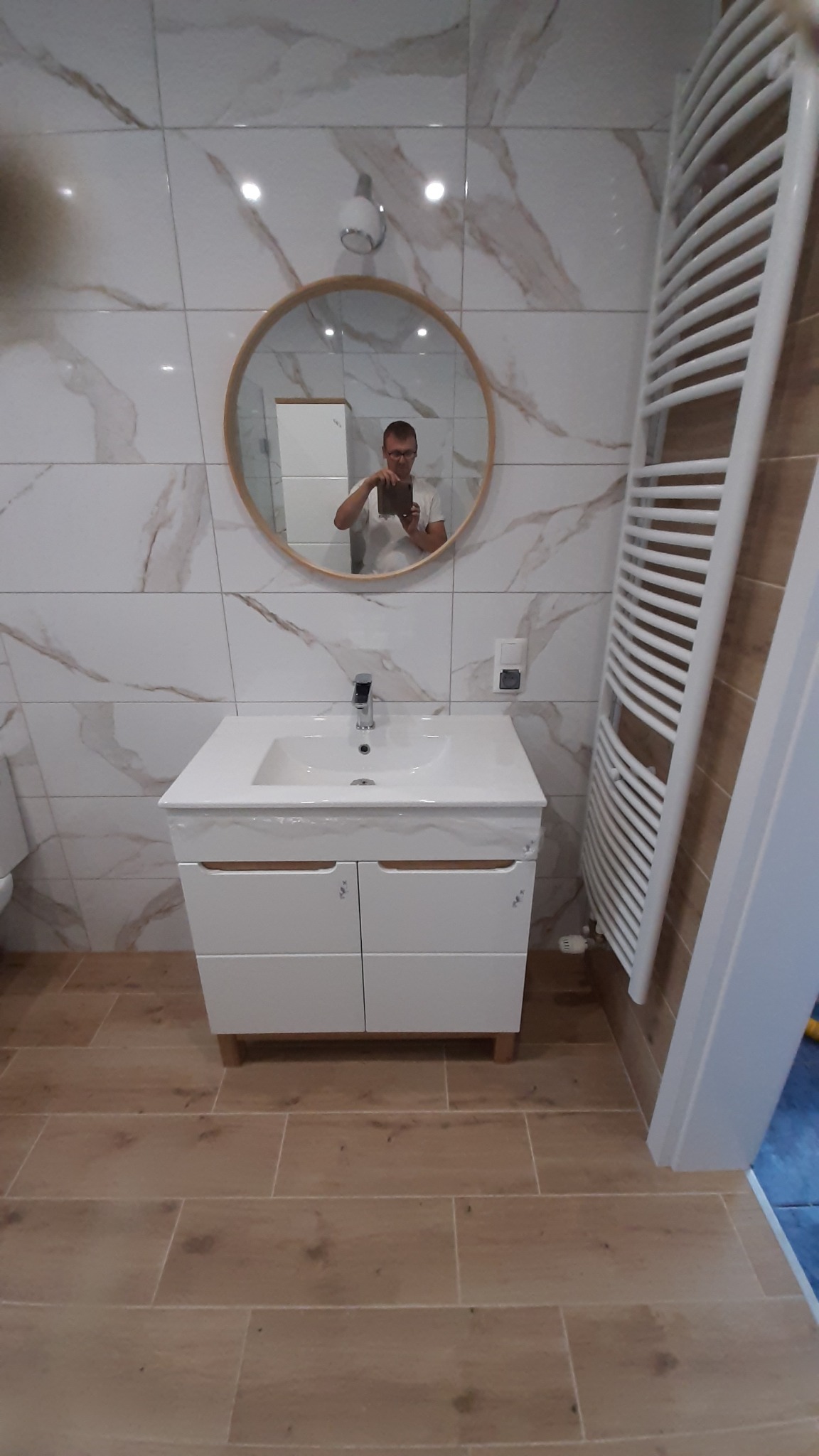 Nowoczesna łazienka z marmurowymi płytkami, okrągłym lustrem z drewnianą ramą i białą szafką pod umywalką, widoczny grzejnik drabinkowy i odbicie osoby robiącej zdjęcie.