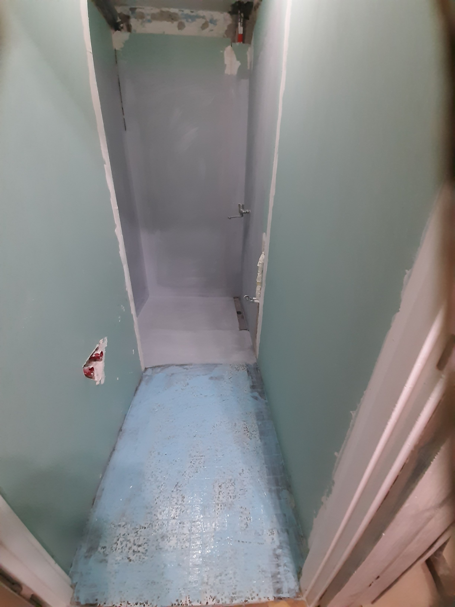 Wnętrze łazienki w trakcie remontu, ściany pokryte płytami kartonowo-gipsowymi, na podłodze pozostałości starej niebieskiej glazury, widoczne instalacje hydrauliczne i elektryczne przed wykończeniem.