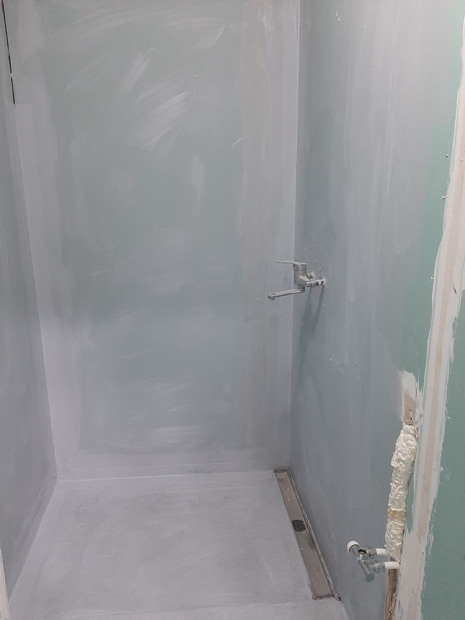 Surowy stan kabiny prysznicowej z widocznym odpływem liniowym, przygotowanej do dalszych prac wykończeniowych. Ściany pokryte szpachlą i wstępnie pomalowane na szaro, z widocznymi rurami i kranem.
