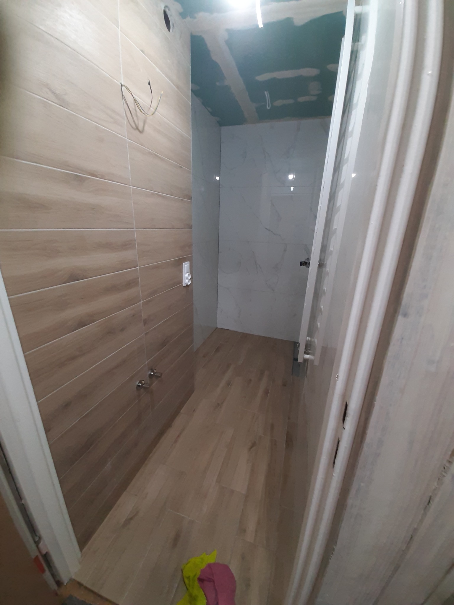 Wnętrze łazienki w trakcie remontu z położonymi beżowymi płytkami imitującymi drewno na ścianach i podłodze, widoczne wystające przewody elektryczne, białe płytki marmurowe w głębi oraz grzejnik...