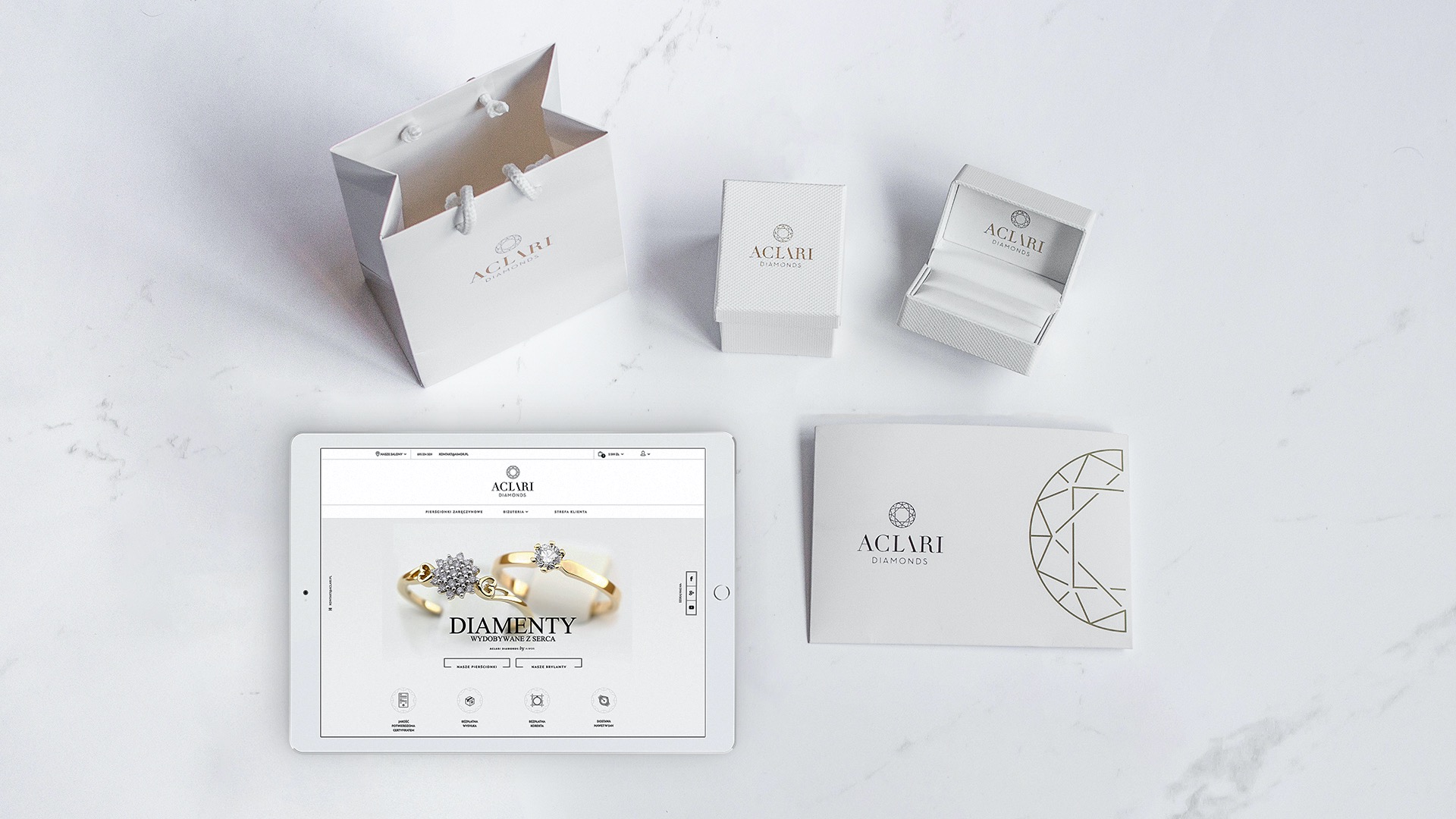 Kompozycja prezentująca identyfikację wizualną marki jubilerskiej: torba, pudełka, broszura i tablet z wyświetloną stroną internetową na marmurowym tle.
