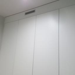 Meble Pod Wymiar - Biała, minimalistyczna szafa wnękowa z ukrytymi zawiasami i kratką wentylacyjną w górnej części, dopasowana do sufitu, widok od dołu.