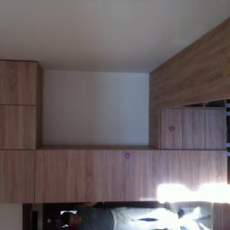 Meble Pod Wymiar - Zabudowa meblowa z drewnopodobnej płyty, widoczny fragment sufitu i odbicie osoby w błyszczącej powierzchni